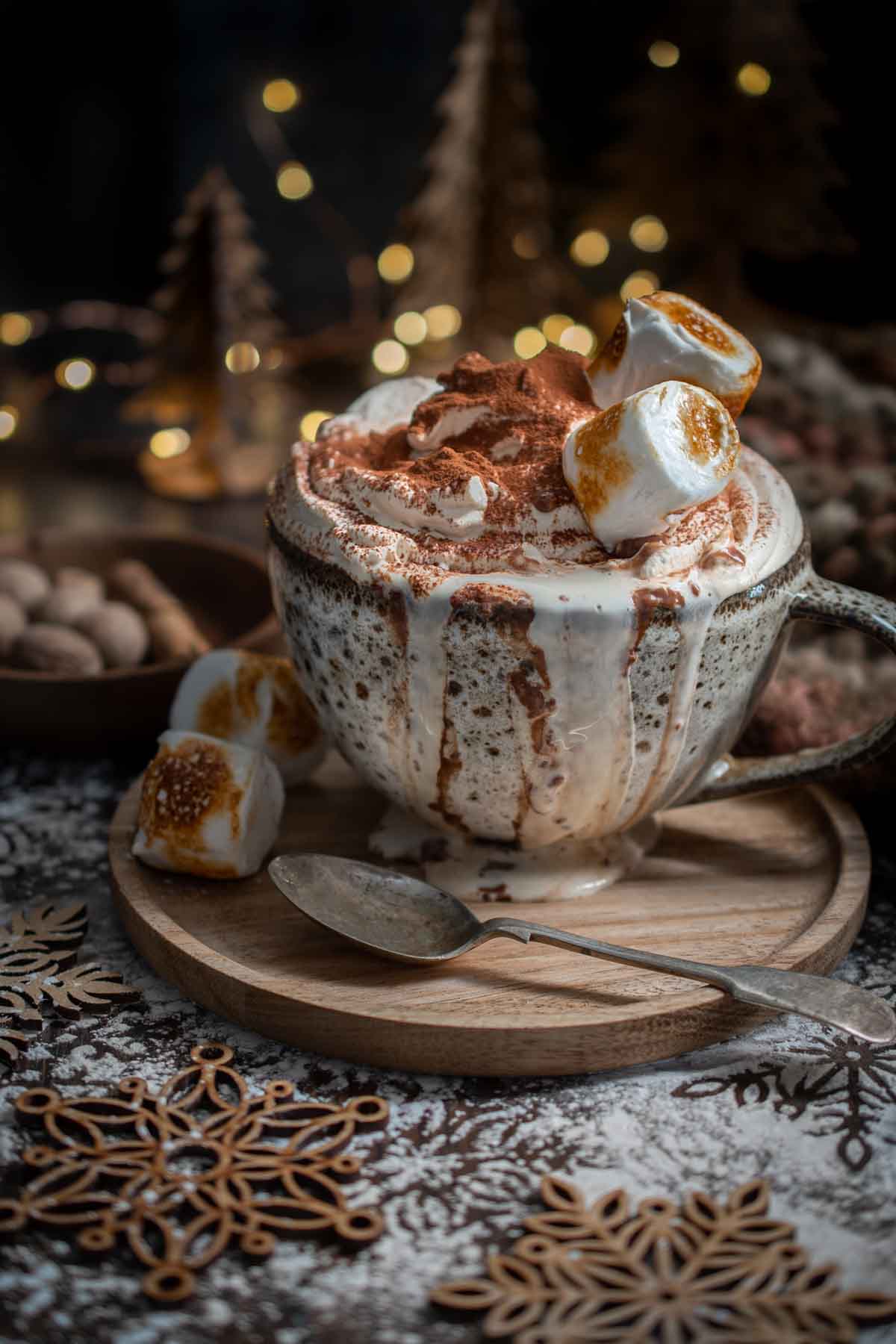 Christmas hot chocolate with cream, toasted marshmallows and cinnamon in a mug with a spoon, festive decorations, and lights