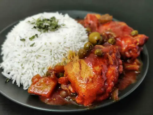 cooked chicken in red tomato sauce with peas served with a rice topped with herbs on the side on a plate