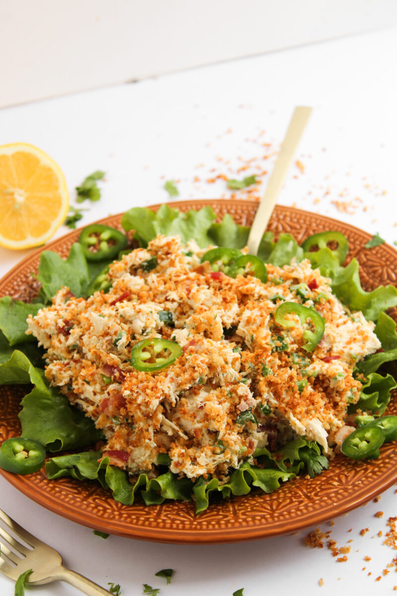 Chicken salad on a bed of lettuce on an orange plate with a sliced lemon and bread crumbs in the background.