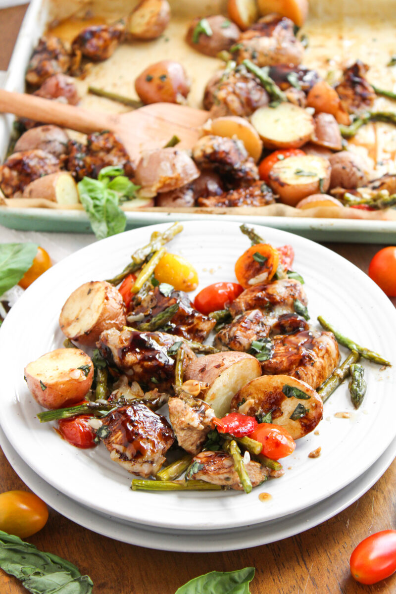 Balsamic chicken and veggies on two stacked white plates with sheet pan in the background.