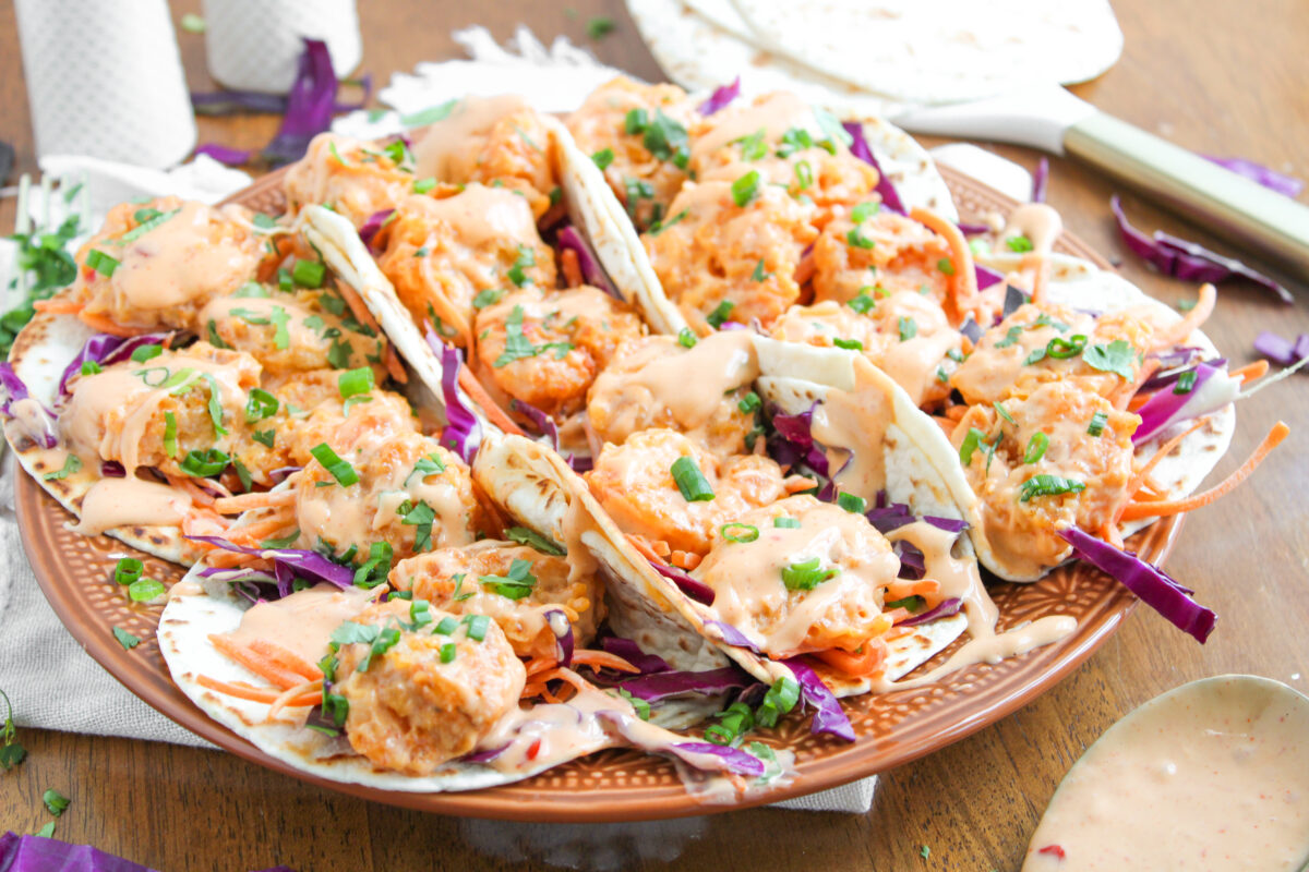 Bang bang shrimp tacos in a row on a large brown plate with tortillas in the background.