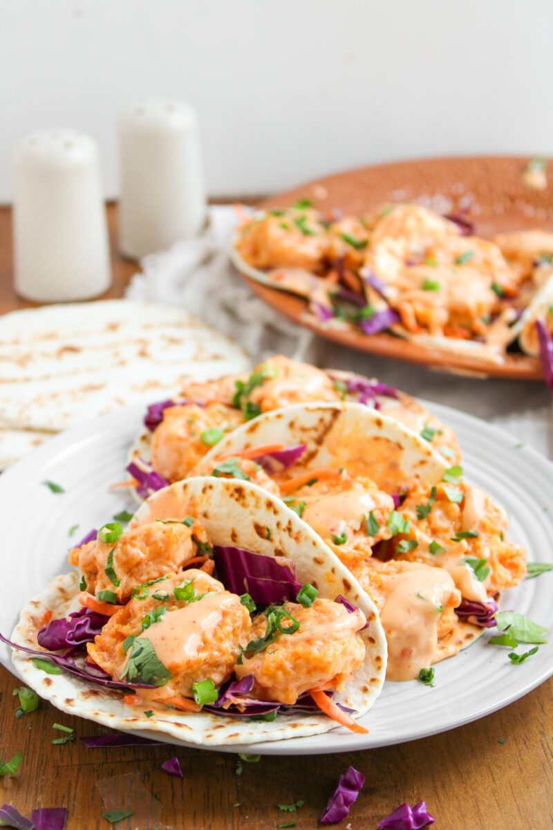 3 bang bang shrimp tacos on a small white plate with more tacos in the background.