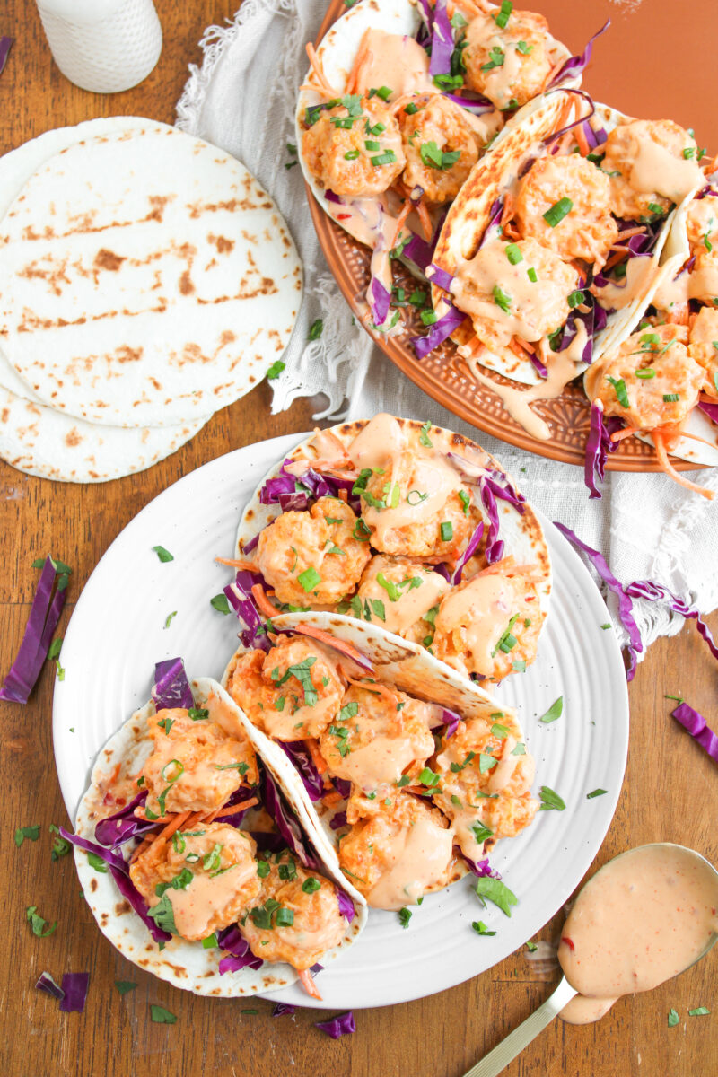 Overhead of 3 bang bang shrimp tacos on a small white plate with more tacos in the background.