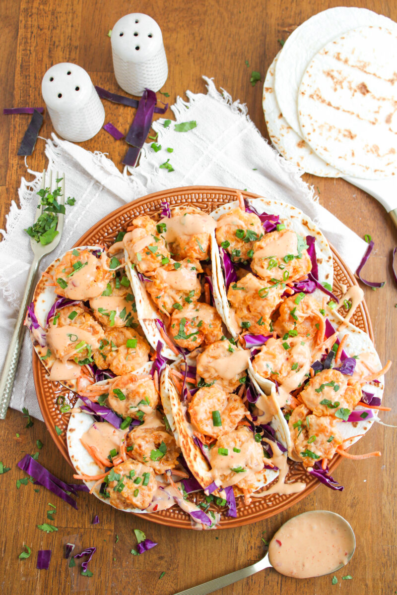 Overhead of bang bang shrimp tacos in a row on a large brown plate with tan napkin underneath.