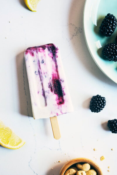blackberry cheesecake popsicle on a white marble countertop with peices of blackberries, lemon wedge and nuts around it