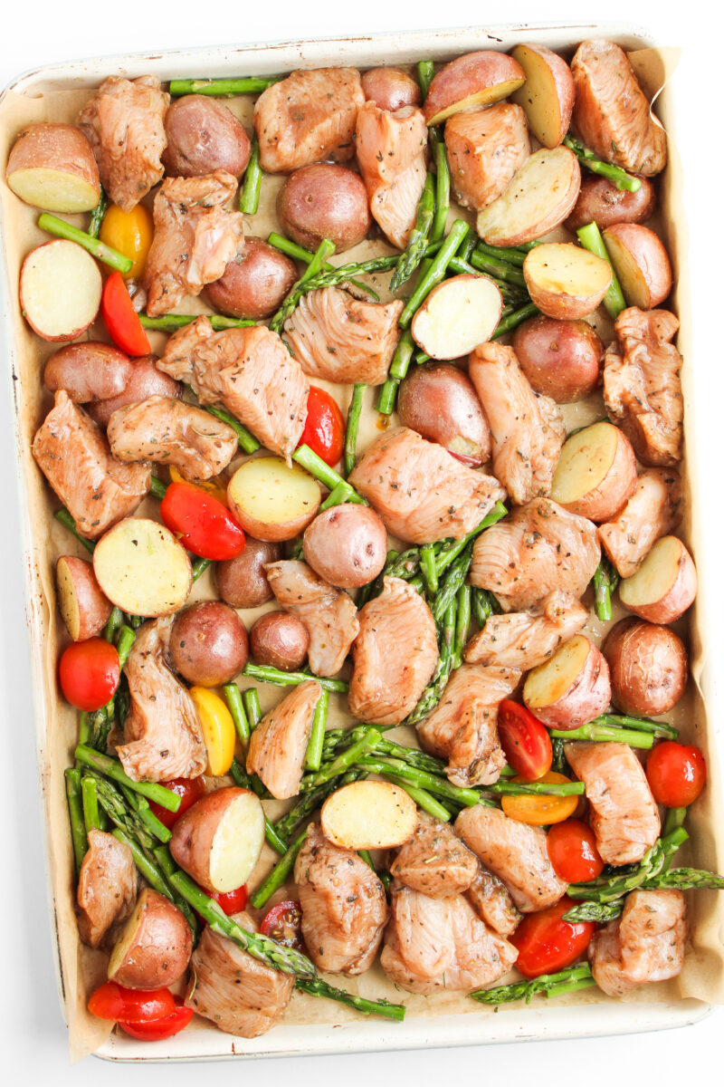 Potatoes, asparagus, tomatoes, and chicken on sheet pan with brown parchment paper.