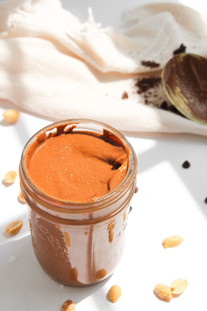 Jar filled with chocolate peanut butter on a white background with sea salt, chocolate chips, and peanuts scattered around.