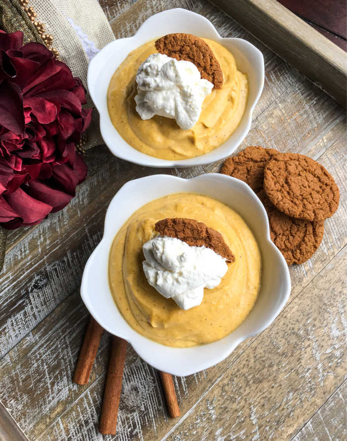 2 servings of creamy pumpkin mousse topped with maple whipped cream, gingersnap cookies, and a sprinkle of ground cinnamon on white serving bowls with cinnamon barks and cookies on the side