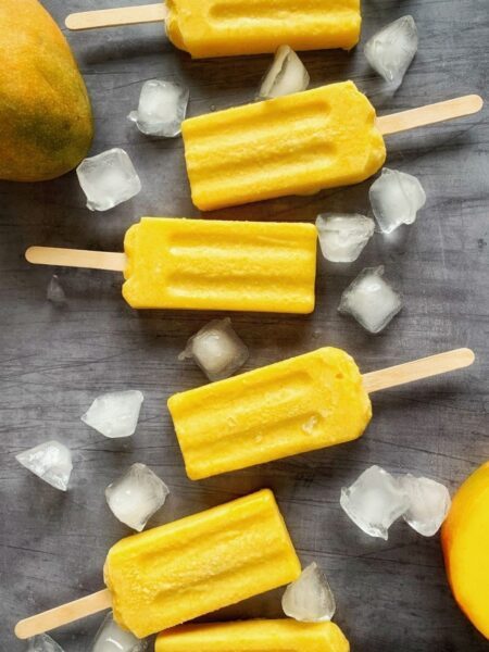 dairy free mango popsicles laid on a gray flat surface with ice cubes and mango fruits