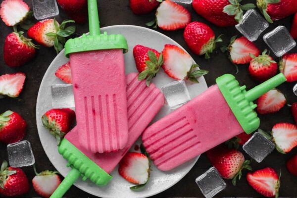 Dairy free creamy strawberry popsicles with sliced straberries and ice cubes on a white plate