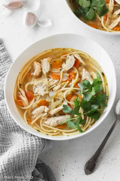 cooked chicken noodle soup with white sliced onions and carrots topped garnished with parsley in a whote bowl with a poon on the side
