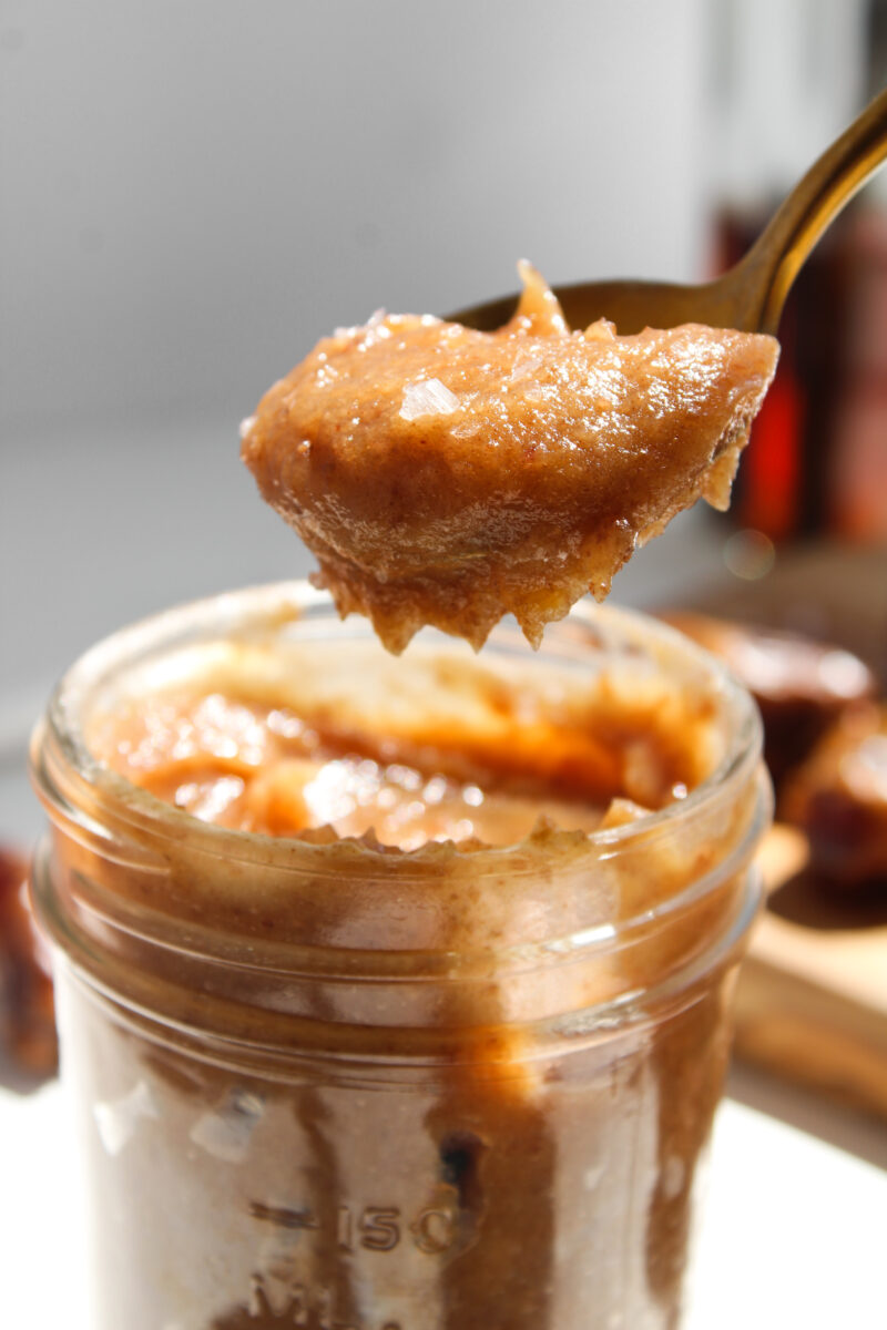 Gold spoon full of date salted caramel lifting to the right of the jar.