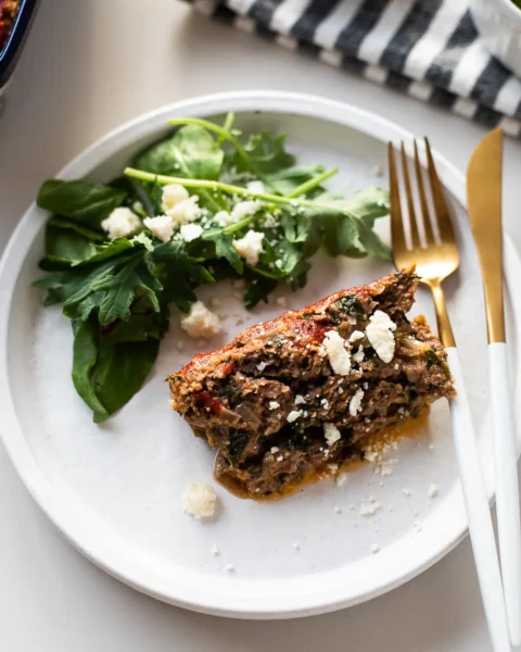a lice of cooked greek lamb meatloaf made with ground lamb, spinach, red onion, oregano, and mint in a white plate