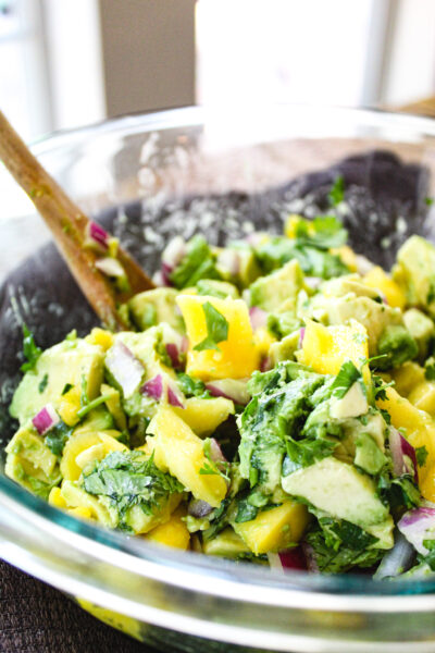 close up view of mango avocado salad in glass bowl and wooden spoon