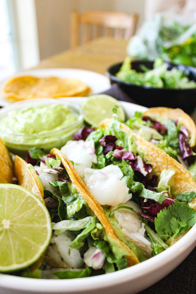 Tacos in a white bowl on a wood table with tortillas, cilantro, and wood chair blurred in the background.
