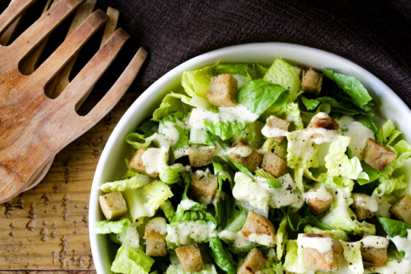 Caesar salad dressing in a white bowl with wooden salad tongs.