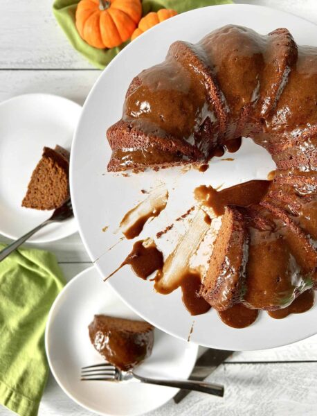 up close image of baked gluten-free Pumpkin Bundt Cake with caramel glaze on a white turntable with saucer plates and forks
