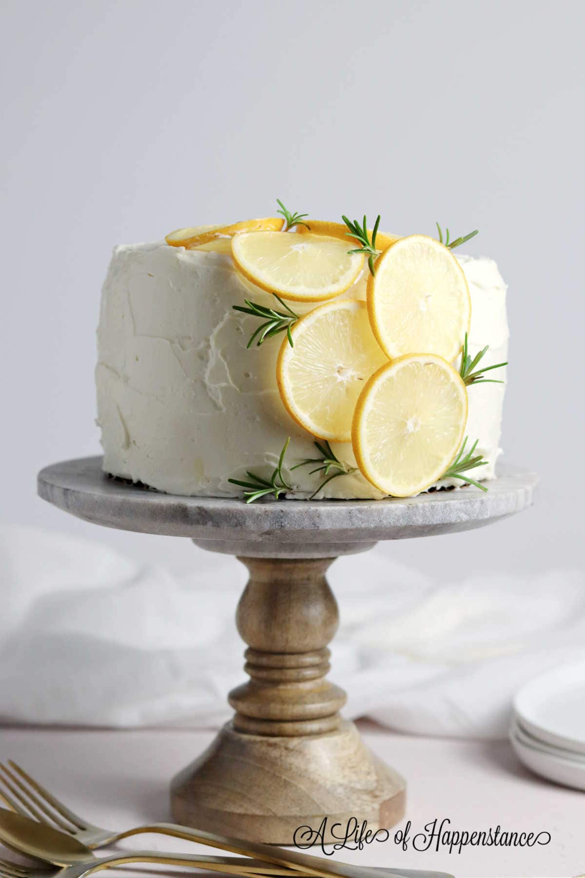 lemon rosemary cake covered with  white cream frosting slices of lemon and rosemary leaves on a wooden cake turntable