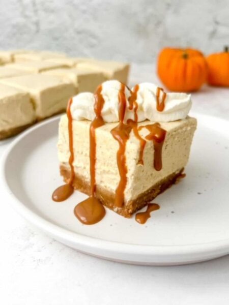a sliced of pumpkin cheesecake bar drizzled with caramel and topped with cream on a white saucer