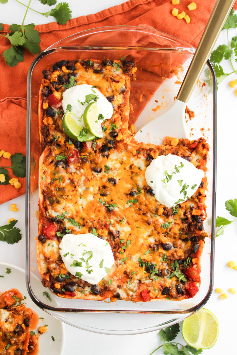 Enchilada casserole in glass casserole dish on white background with orange napkin and ingredients scattered around.