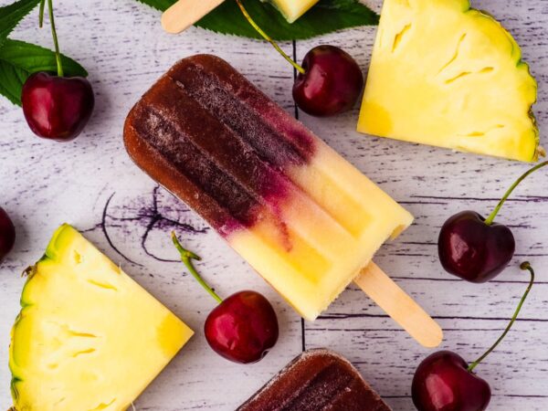 pineapple cherry popsicles on a white wooden table with cherries and slices of pineapple 