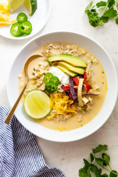 creamy chicken chili topped with  slices of avocadoes, jalapeño parmesan, a dash of sour cream  and a slice of lime served in a white bowl with a golden spoon