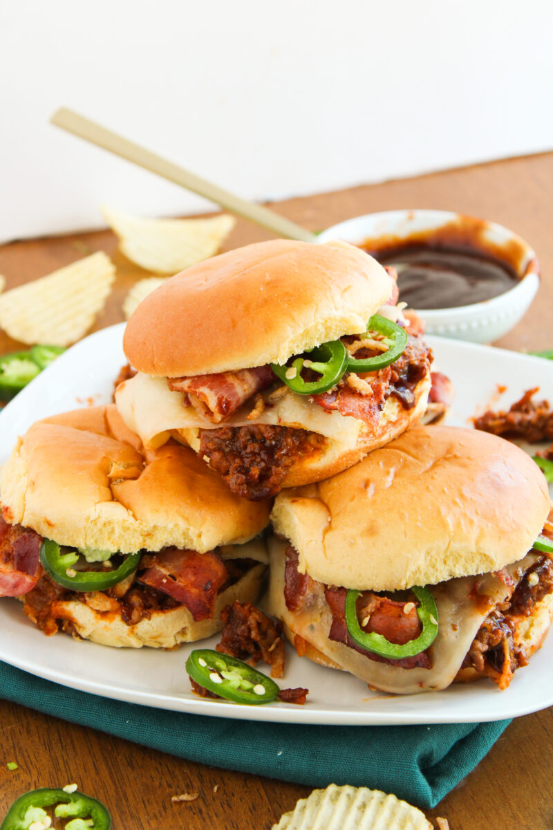 Angled view of three sloppy joes in a pile on a white plate with a green napkin underneath on a wood background.