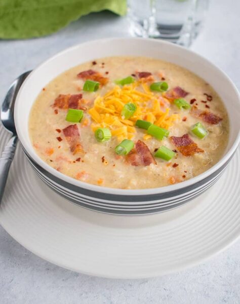 cauliflower cheese soup topped with slices of cheese, bacon and chives served in a white bowl with spoon on the side