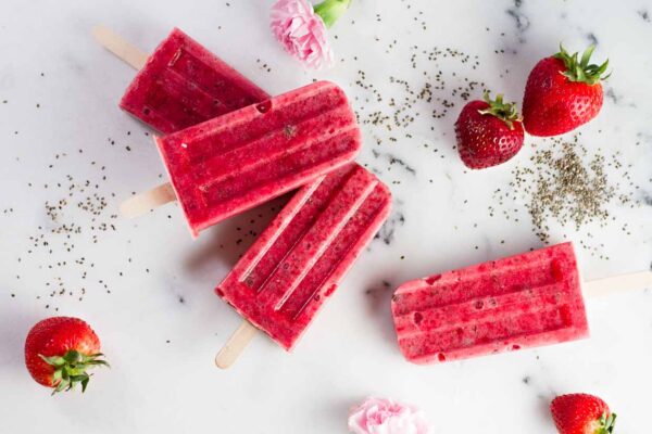 dairy free strawberry chia seeds popsicles on a white flat surface with strawberry fruits on the side, pink flowers, and a sprinkling of chia seeds 