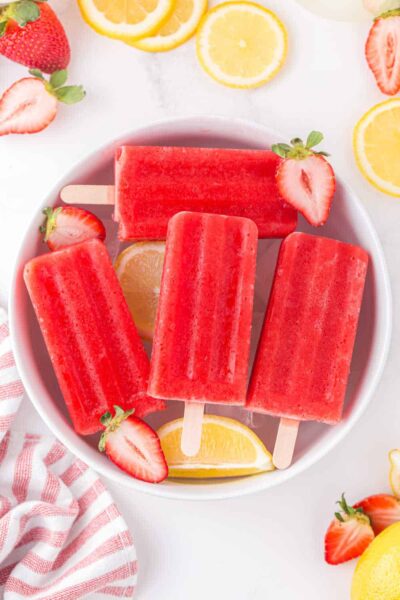 strawberry lemonade popsicle served on top of ice cubes with slices of strawberry and lemon in a white bowl on a white countertop