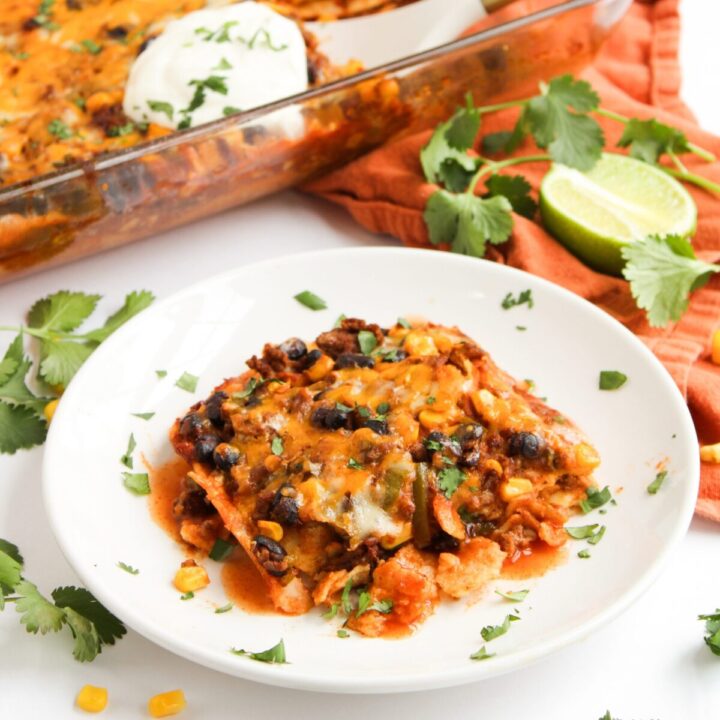 Serving of beef enchilada casserole on a white plate with casserole dish in the background.