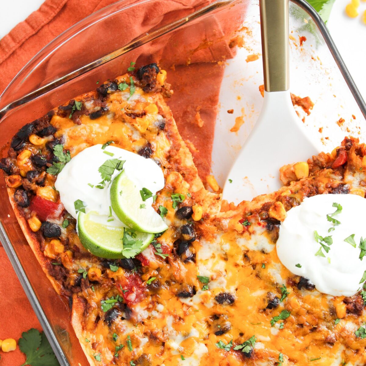 Beef enchilada casserole dish at an angle with white and gold serving spoon tucked in on an orange napkin.