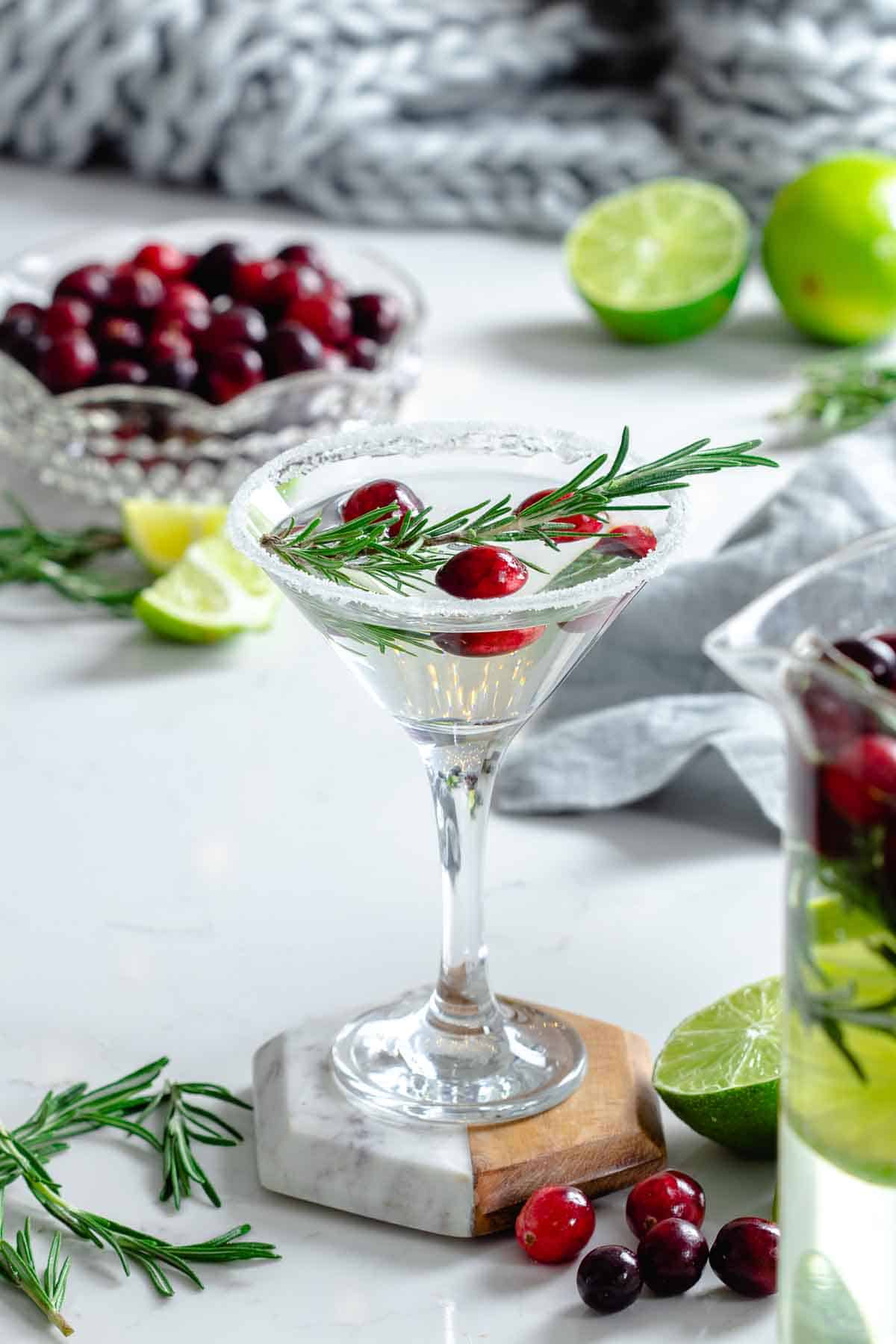 white cocktail granished with cranberries and rosemary leaf in a martini glass, with lime, cranbeeries and rosemary leaves on the background