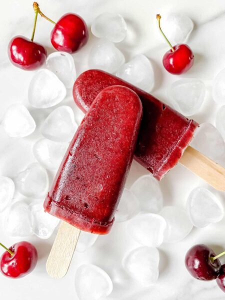cherry mango popsicles with ice cubes with cherries on a white flat surface
