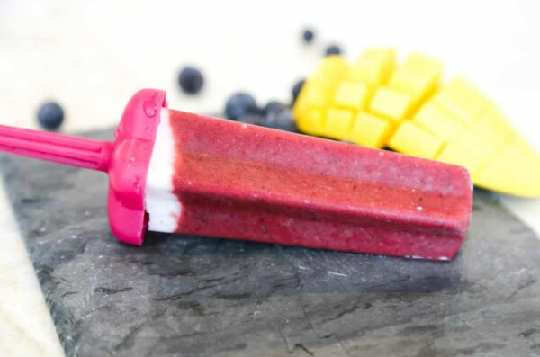 a piece pf cherry popsile on a gray black surface with slice cubes of mango and blueberries on the side