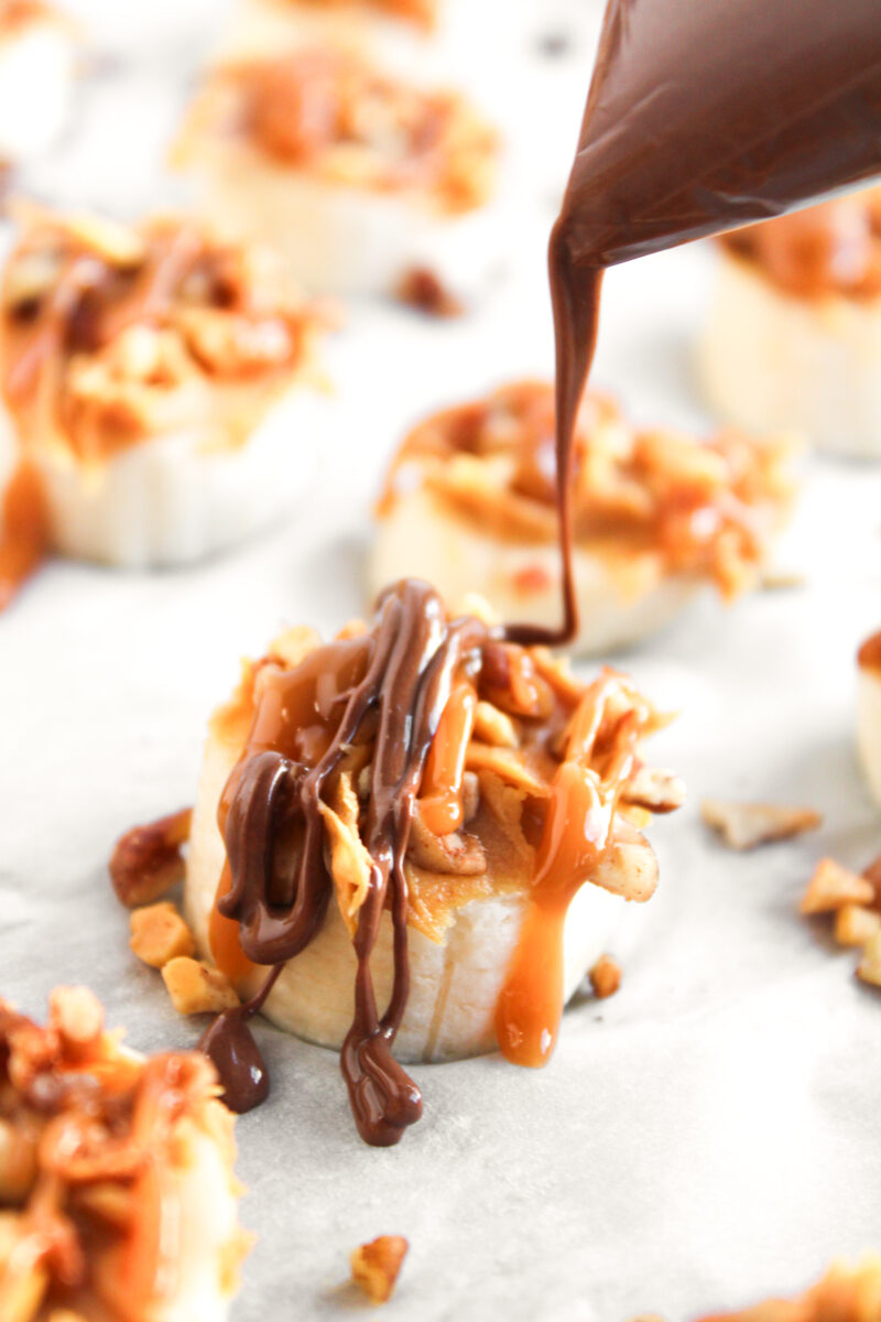 Melted chocolate being drizzled over banana slice on white parchment paper