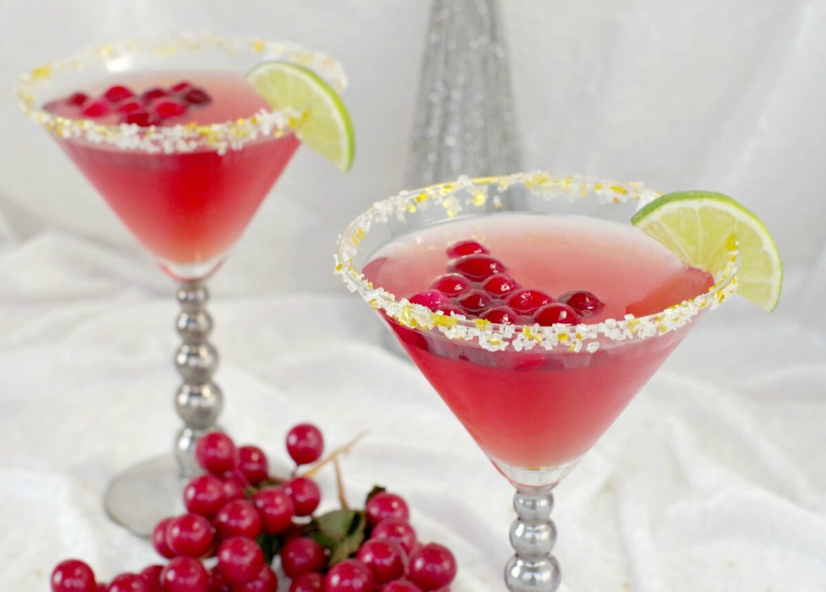 festive cranberry cosmo mocktail with cranberries and lime wedge in small cocktail glasses rimmed with white sugar and edible glitters