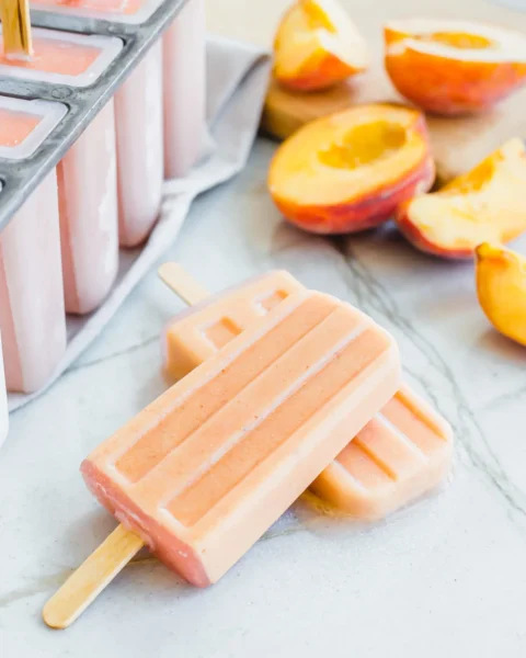 2 pcs dairy free peach popsicles on white granite marble countertop with slices of peach fruit
