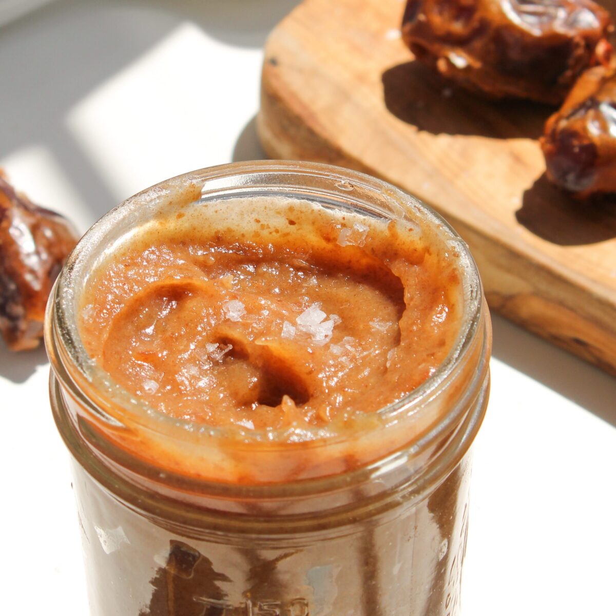 Date caramel in glass jar with wood cutting board behind it.