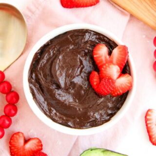 Overhead of chocolate avocado pudding in white bowl with heart shaped strawberries surrounded by pink napkin, wood board, and avocados.