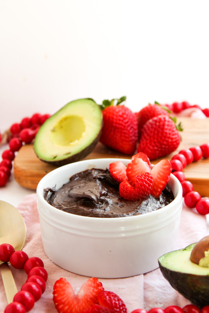 Chocolate pudding in white bowl with heart strawberries, avocado, and pink napkin in background.