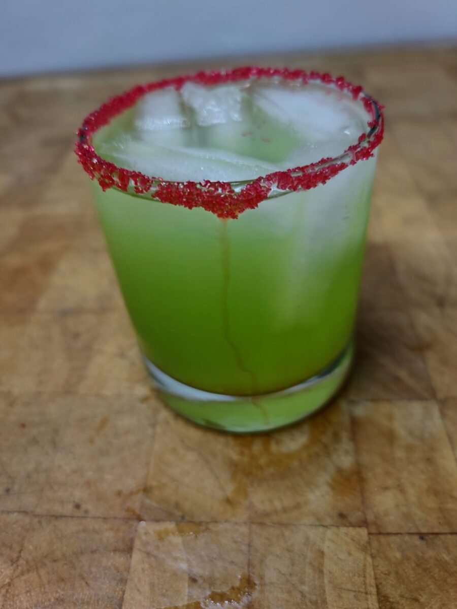 green colored boozy grinch on a glass with ice rimmed with red sugar 
