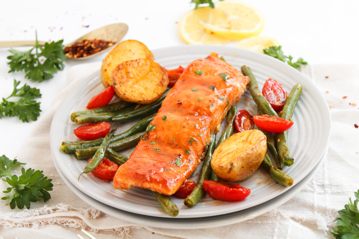 Salmon and vegetables on a serving plate surrounded by a napkin, parsley, lemon, and a gold spoon holding crushed red pepper.