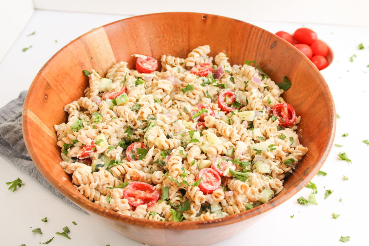 Tuna pasta salad in wood bowl with parsley scattered around, blue napkin, and red tomatoes in a bowl.