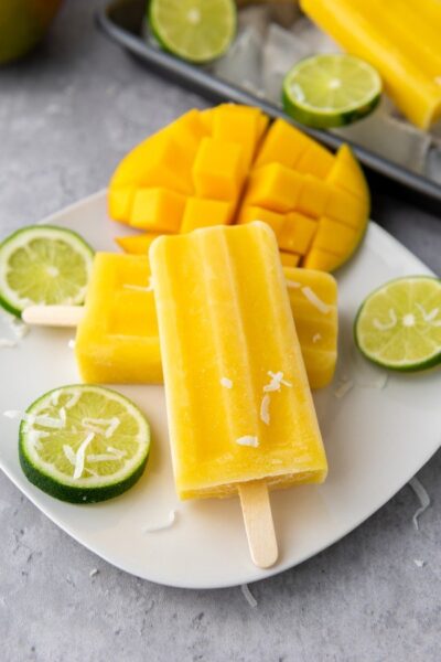 mango lime popsicles on a small square white plate  with slice cubed mango fruit and slices of lime and a sprinkling of shredded coconut meat
