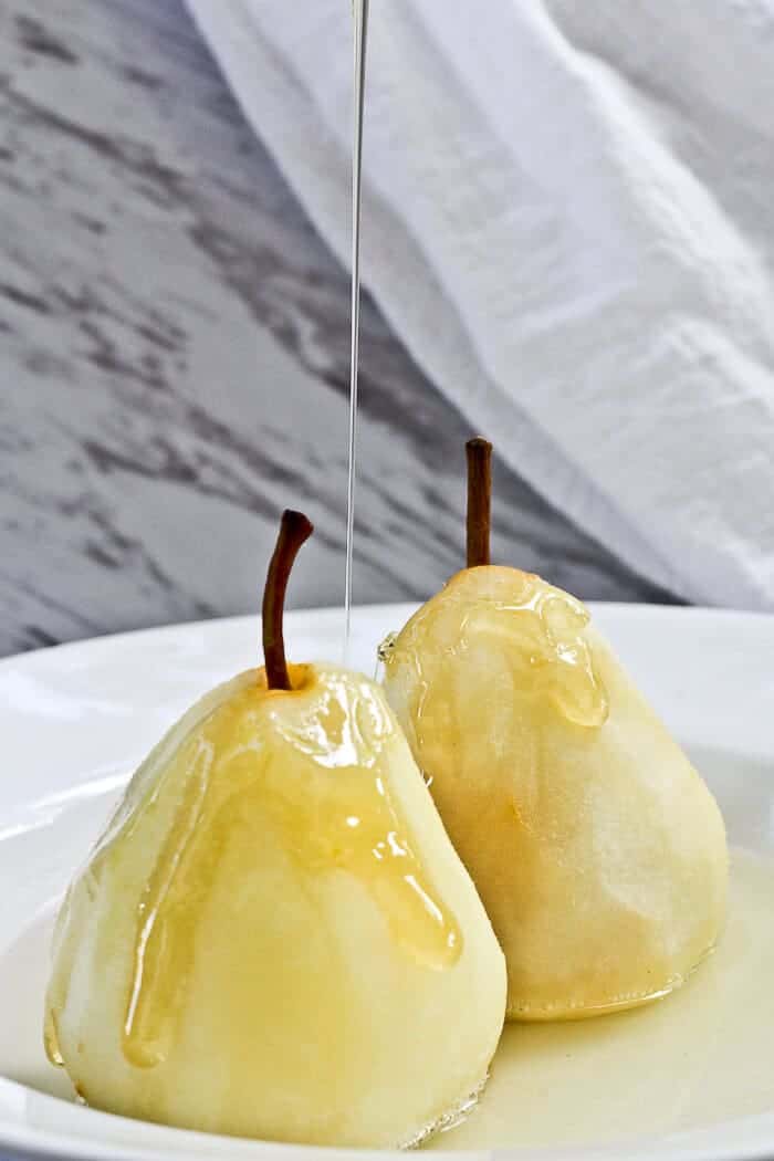 2 white poached pears deing drizzled with syrup on a white deep plate