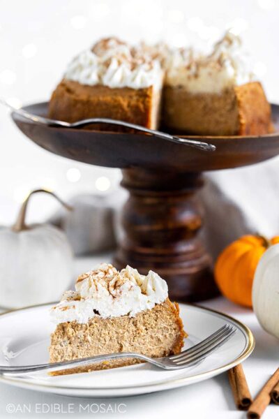 a slice of keto pumpkin cheesecake topped with swirls of cheesecake sprinkled with cinnamon on a white saucer plate with a fork,  in the background are the rest of the  pumpkin cheesecake on a cake turntable, pumpkins, and cinnamon sticks