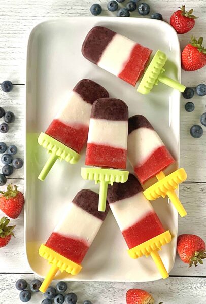 Red, White and Blue Paleo Popsicles on a white rectangular plate on a white wooden table with straberries and blueberries on the table`