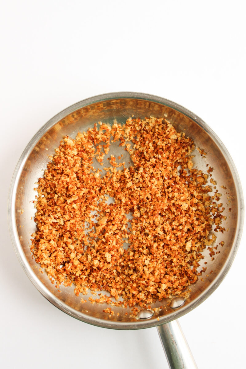 Toasted breadcrumbs in a silver sauce pan on white background.