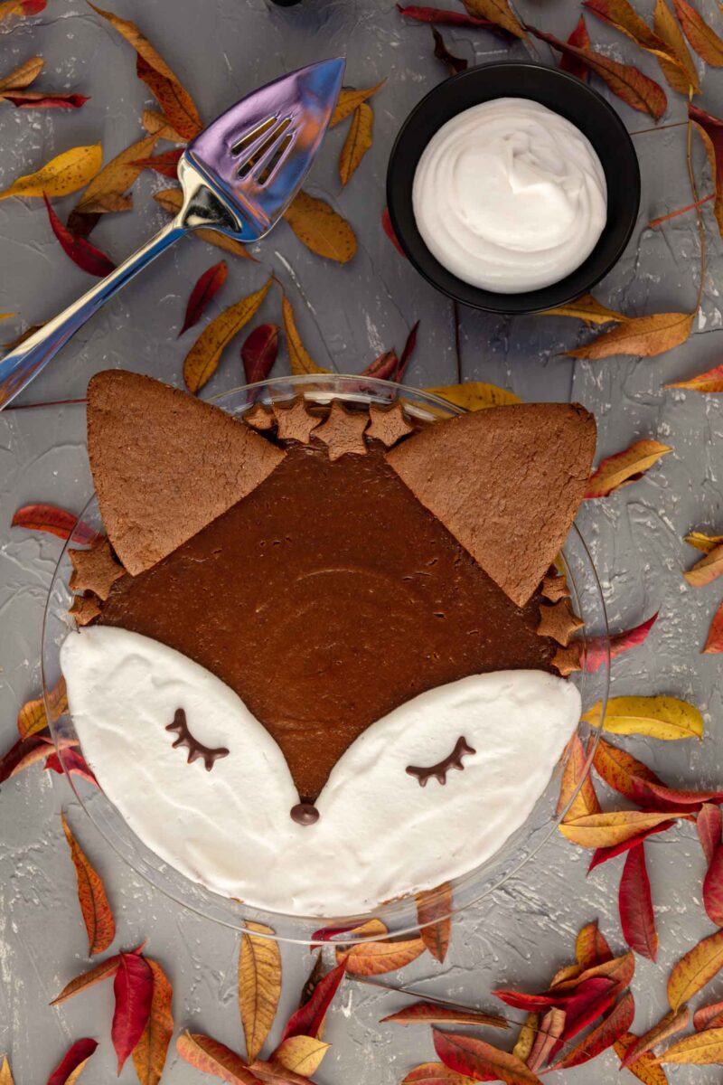 baked vegan crustless pumpkin pie decorated as a fox , in the backgorund- maples leaves and a cream in a glass sauce bowl